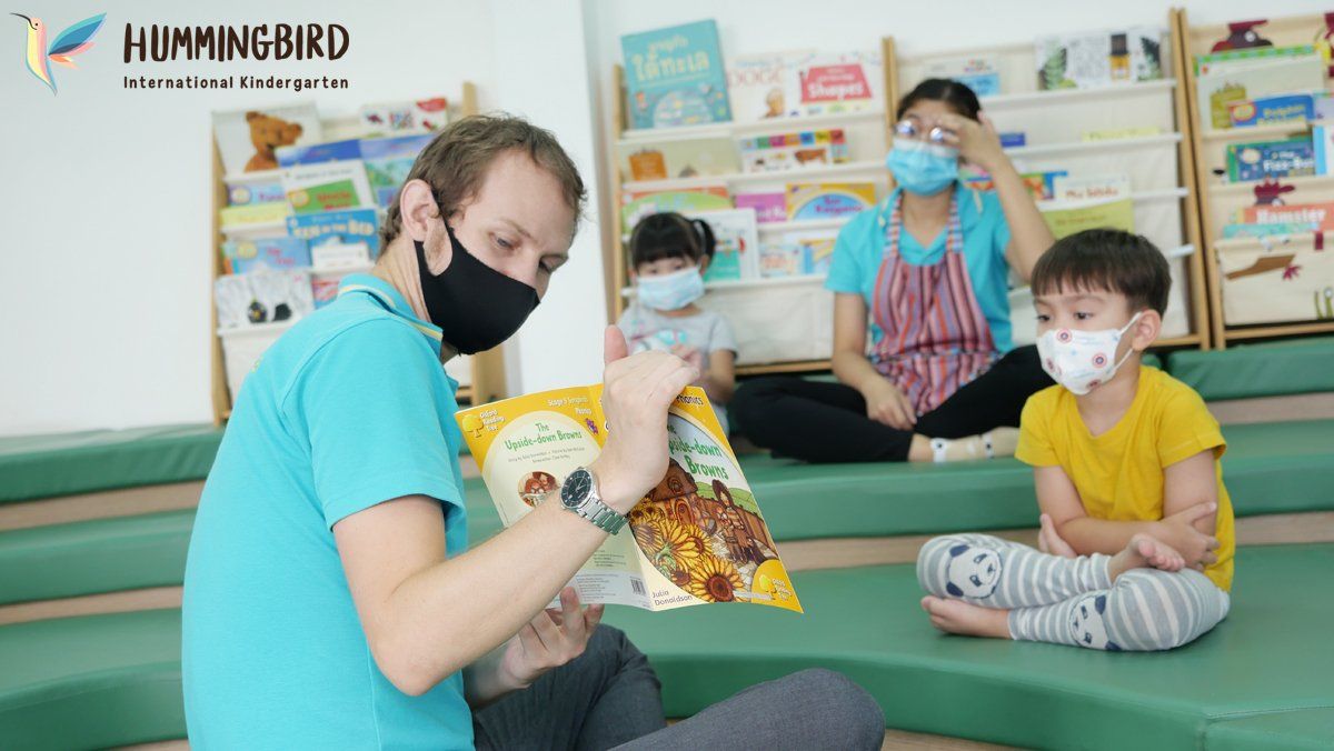 A man wearing a mask is reading a book to a group of children.