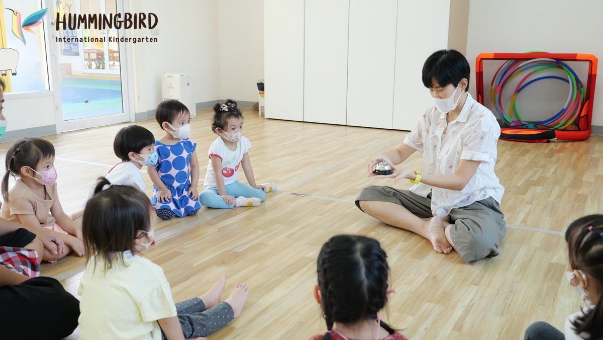 A group of children are sitting on the floor in a room with a man wearing a mask.