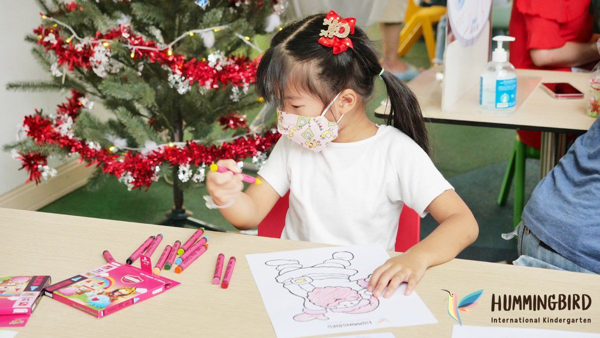A little girl wearing a mask is sitting at a table with a christmas tree in the background.