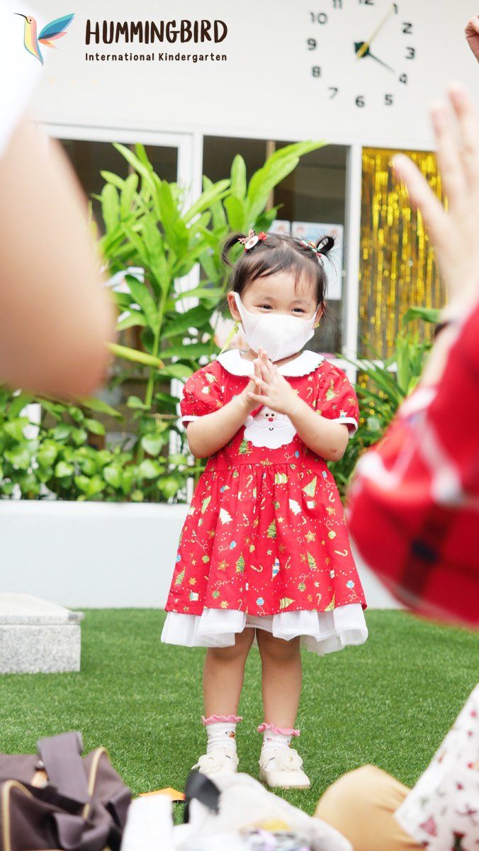 A little girl wearing a red dress and a mask is clapping her hands.