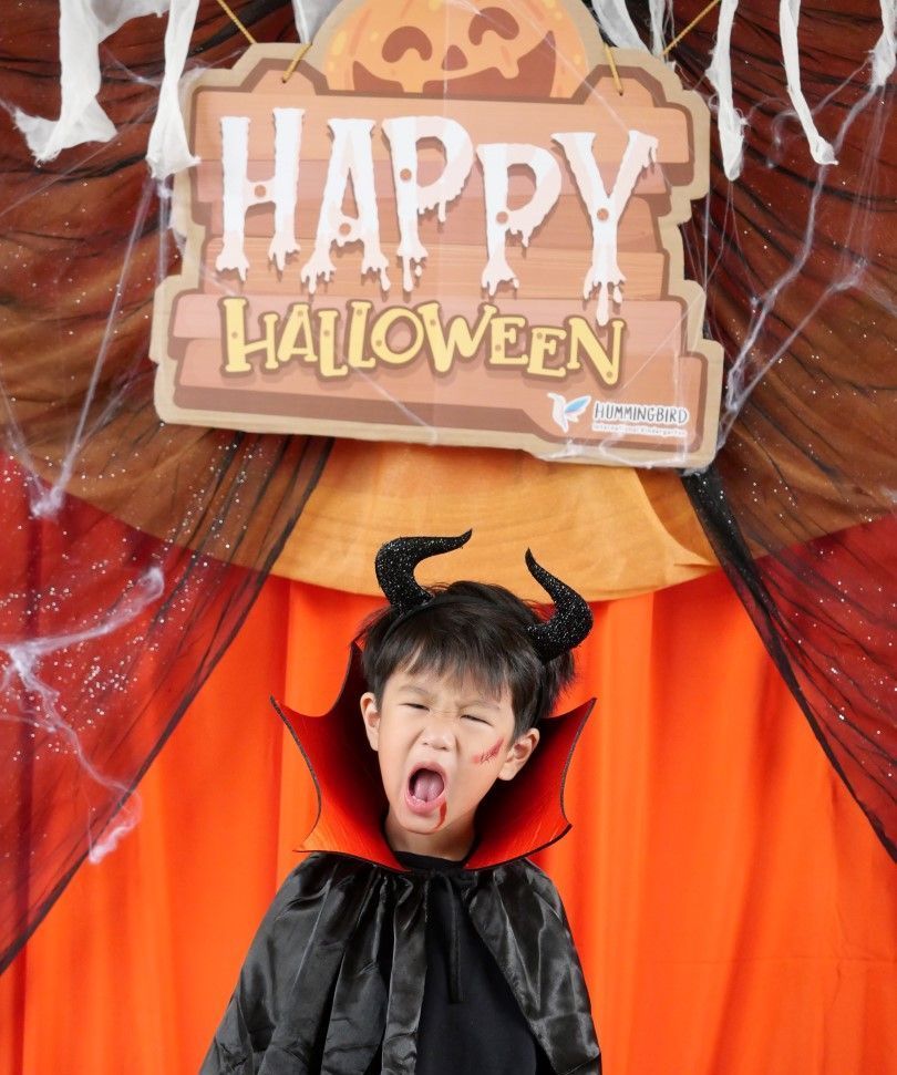 A little boy in a devil costume stands in front of a happy halloween sign
