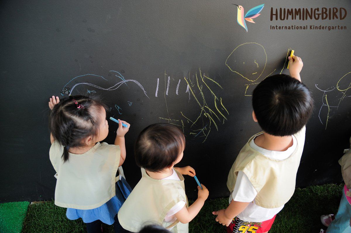 Three children are drawing on a blackboard in front of a hummingbird logo