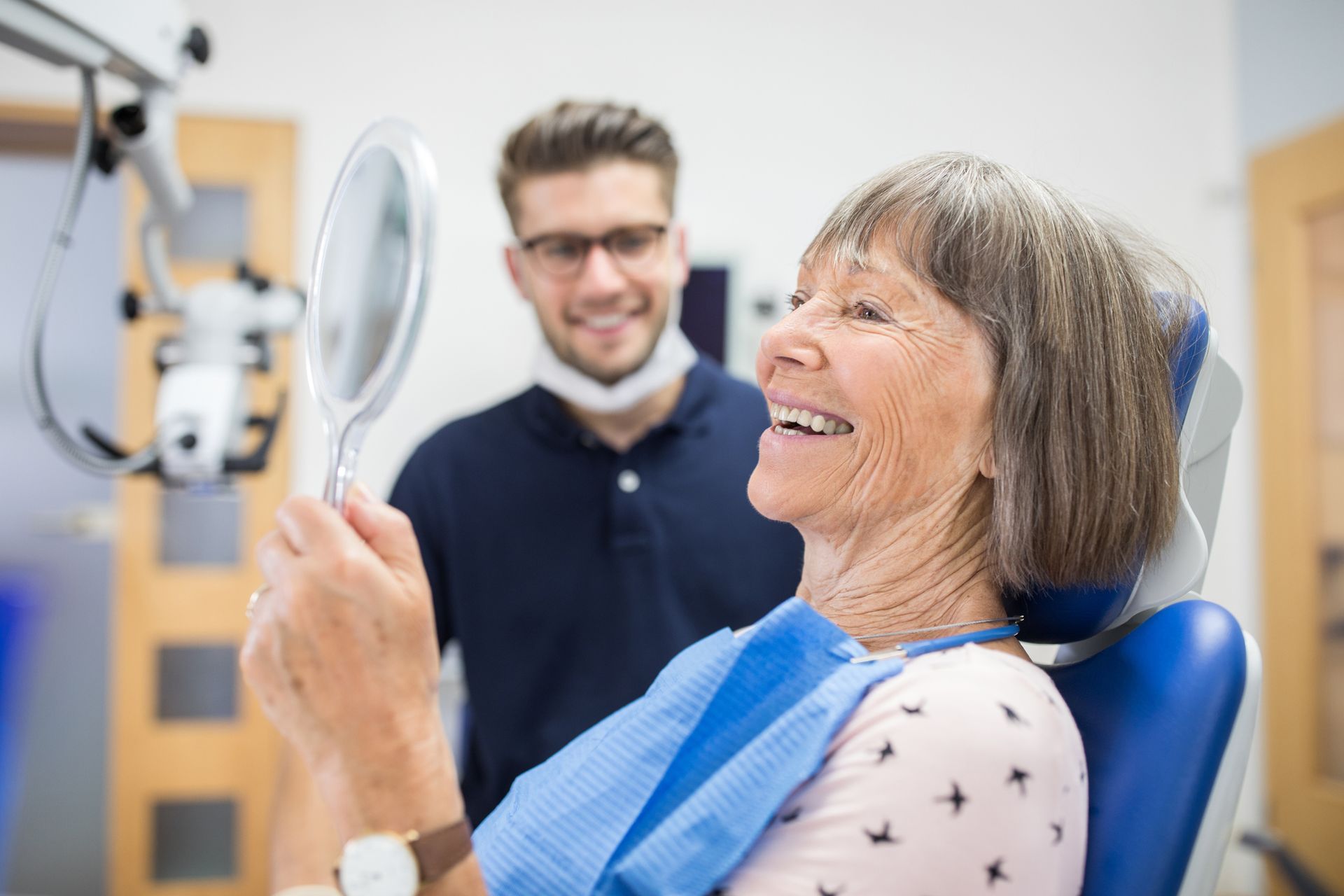 An elderly woman is sitting in a dental chair looking at her teeth in a mirror.