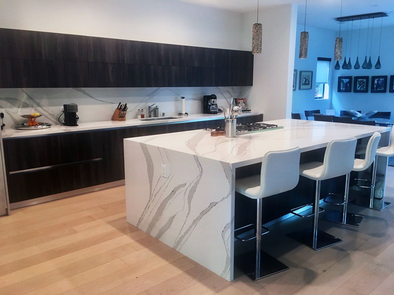 Modern kitchen with large island and bar seating. Dark wood cabinets, white marble countertops, and blonde wood floors.