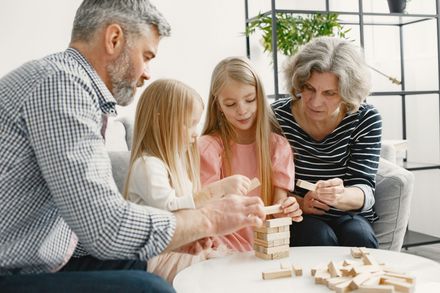 Family building a wooden block tower at a table, with a man, two children, and an older woman nearby.