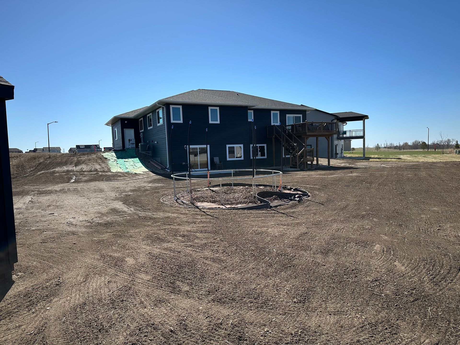 Two-story blue house with a deck and fire pit on a gravel lot under a blue sky.