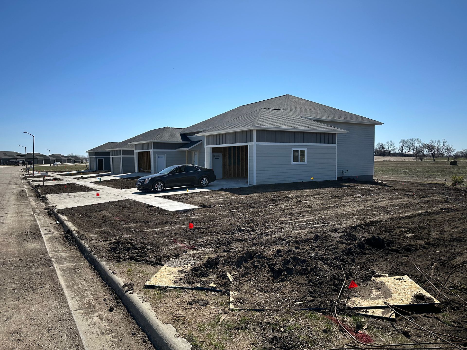 New construction houses with open garages, blue siding, and dirt yards, under a clear, sunny sky.