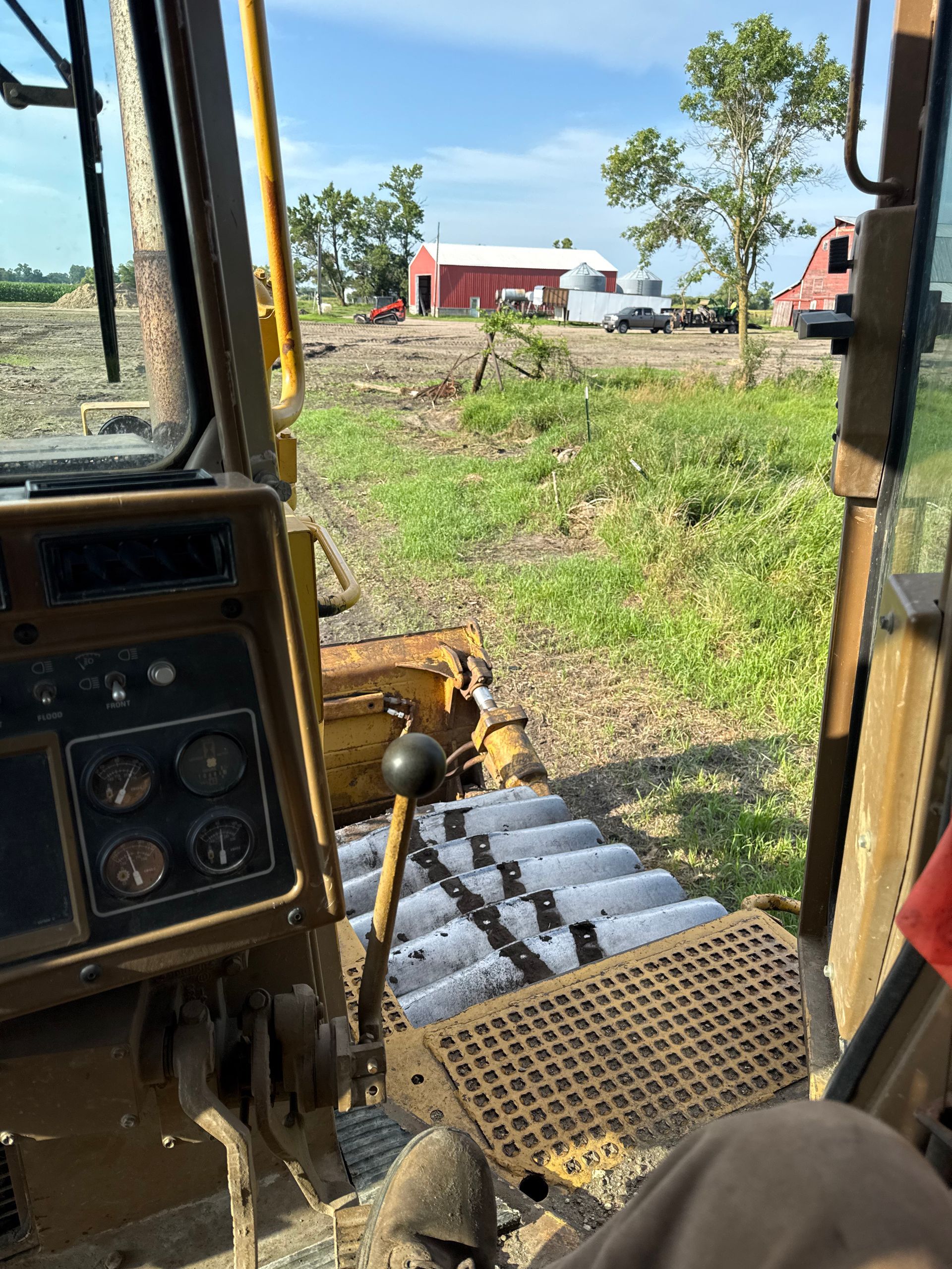View from inside a bulldozer cab, looking out at a field and red barns on a sunny day.