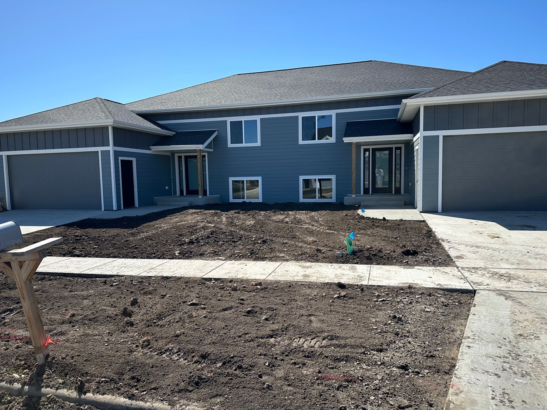 Two blue duplex units with gray garage doors and a bare yard under a blue sky.