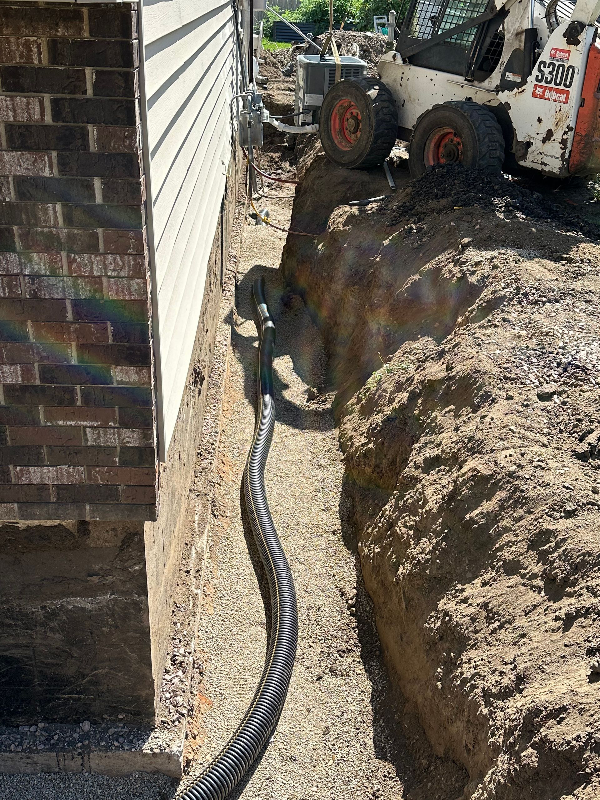 Trench along a house foundation with black drainage pipe, dirt, and a skid steer.