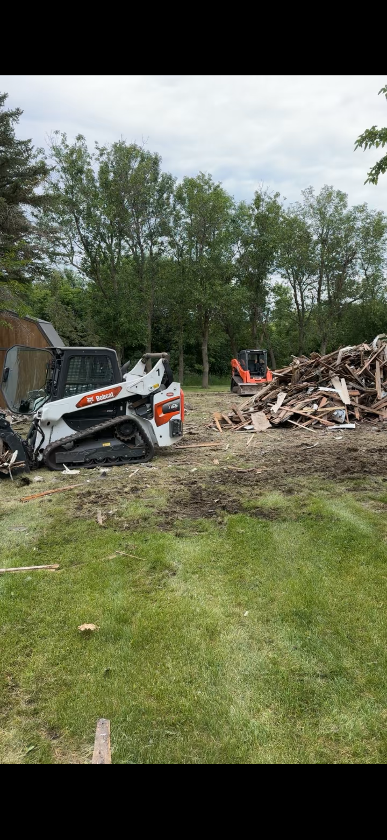A white Bobcat machine, other machinery, and wood debris on a grassy area in front of trees.