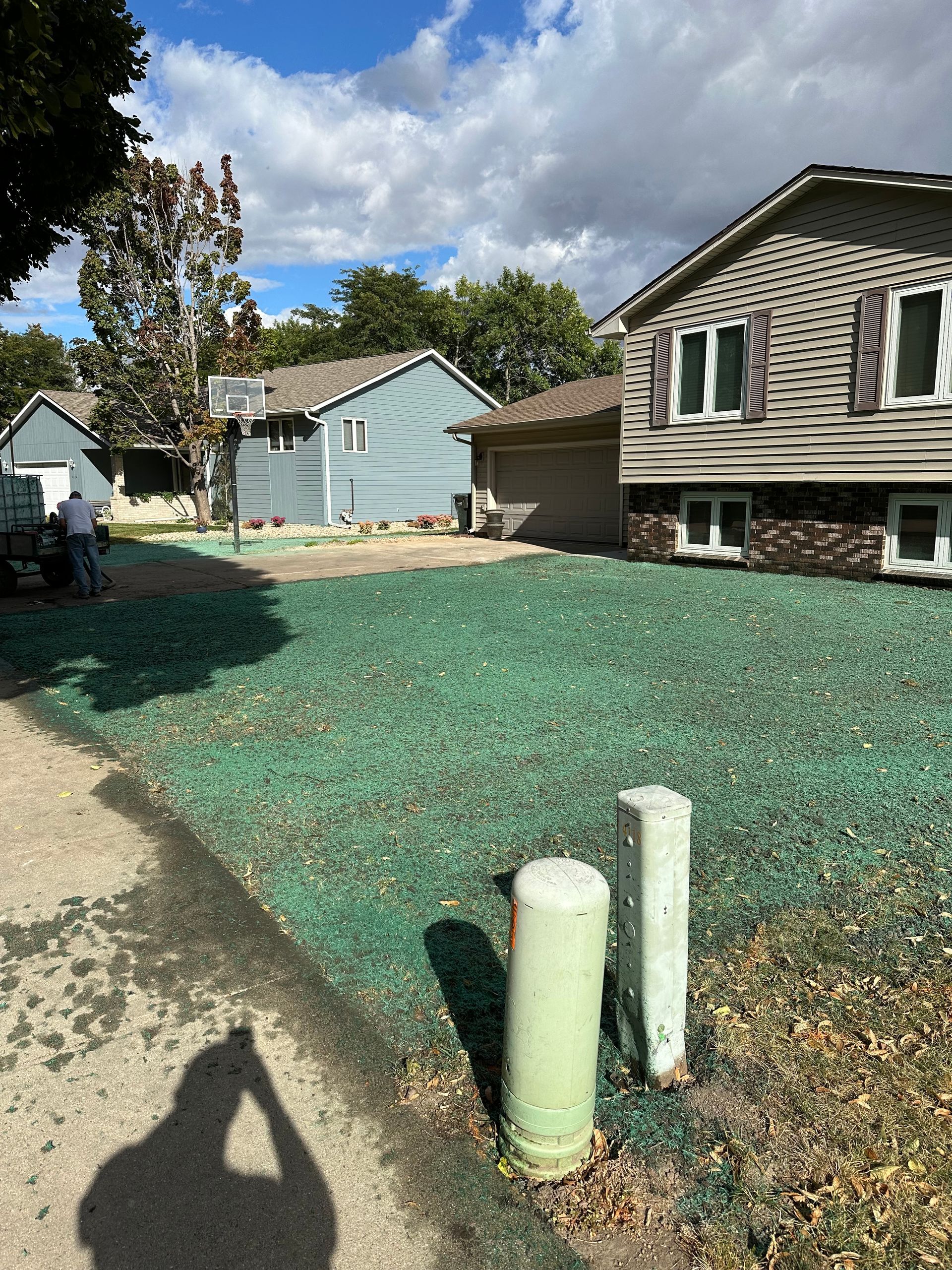 Green-covered lawn in front of houses; street view. A person works on the lawn. Cloudy, blue sky.