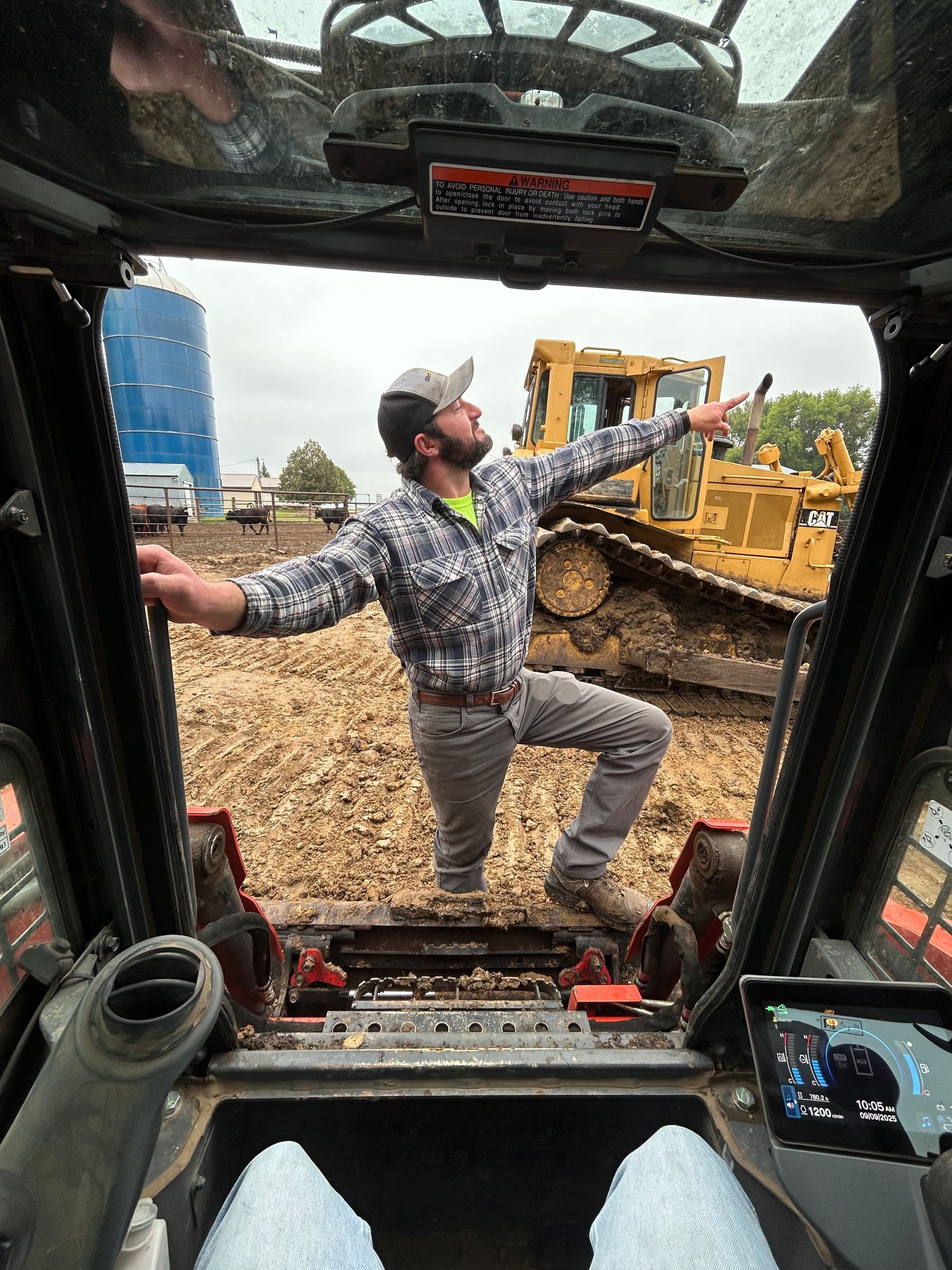 Man in plaid shirt and work boots stands arms outstretched near a bulldozer. Viewed from inside a construction vehicle. Muddy setting.