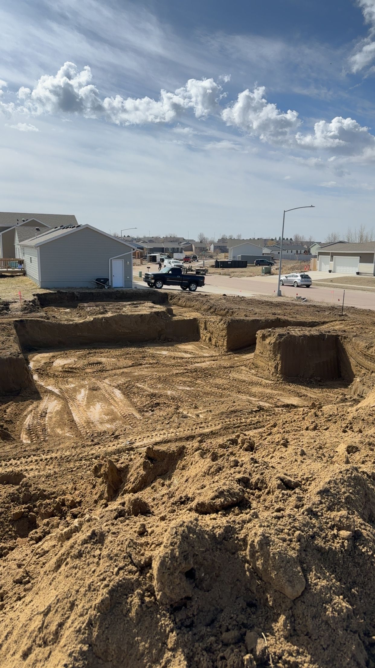 Construction site with excavated foundation. Earth mounds in foreground, houses in background under cloudy sky.