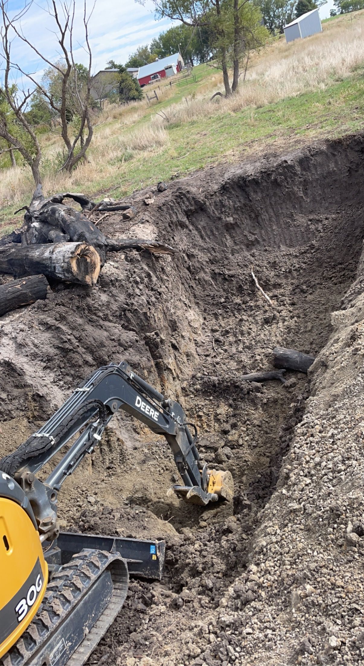 Mini-excavator digging a trench in a brown earthen slope.  A pile of black fabric and vegetation rests along the edge.