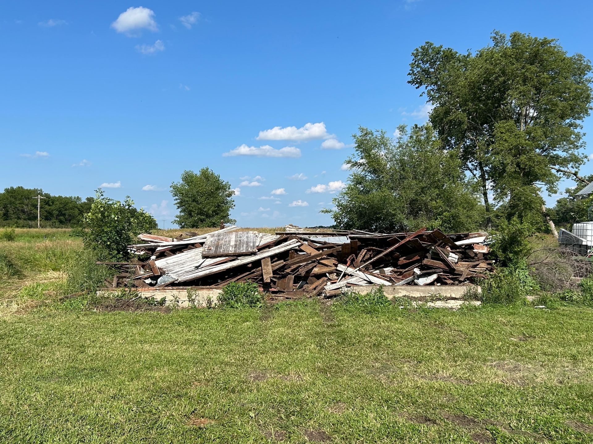 Pile of wood debris on a concrete foundation in a grassy field under a blue sky.