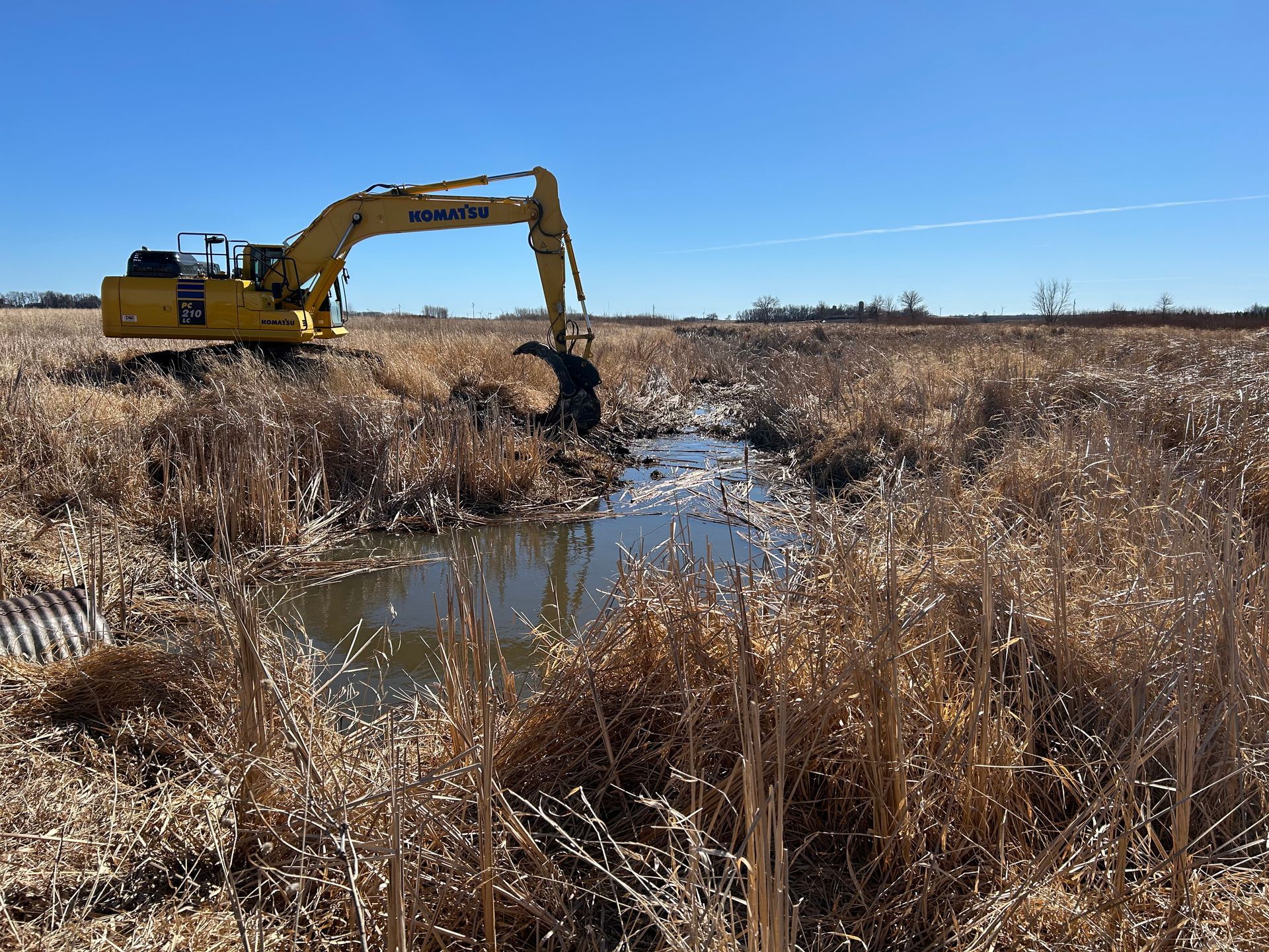 A yellow excavator clearing a water channel through tall, dry grass on a sunny day.