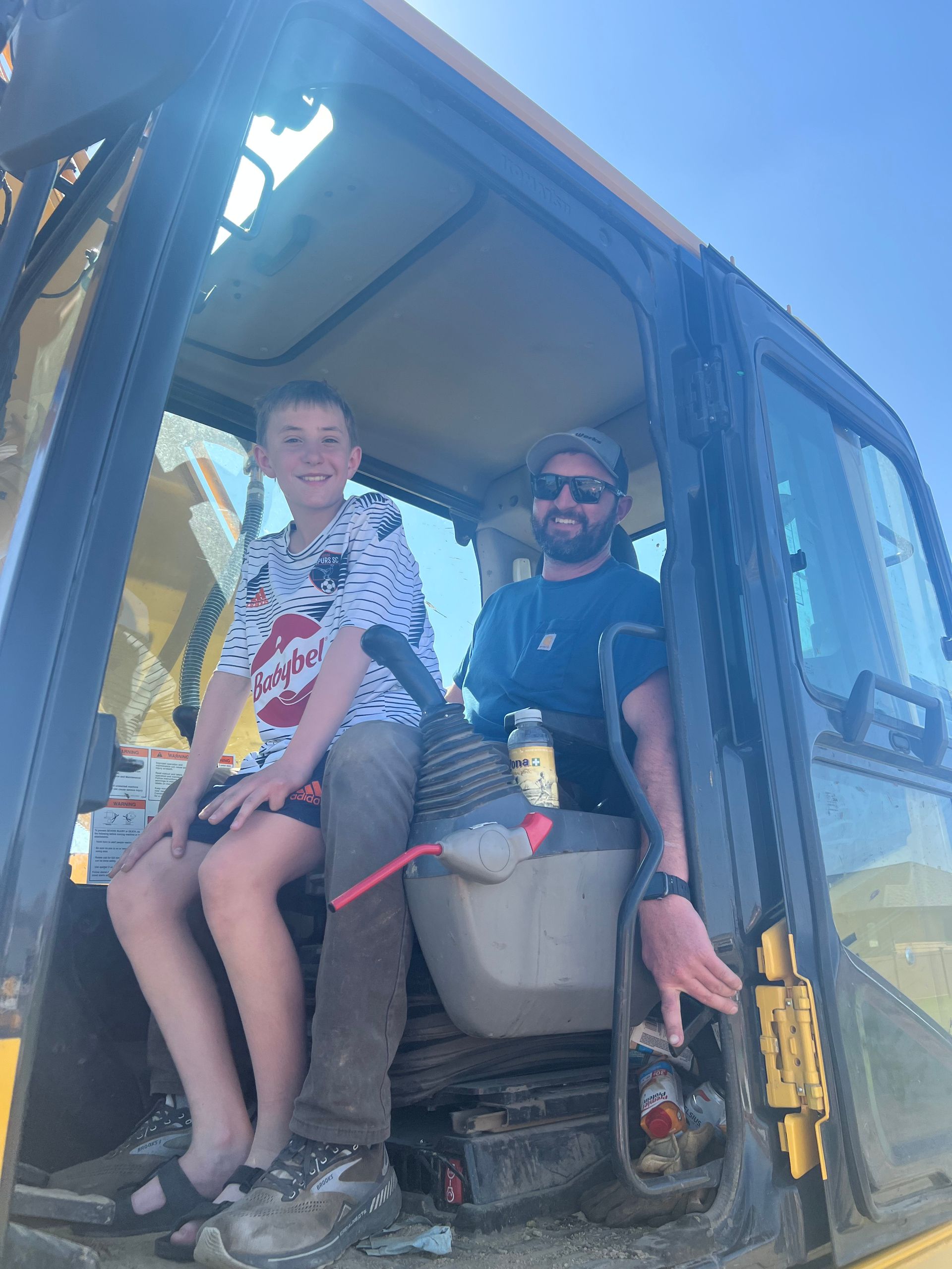 Man and child sit in a construction vehicle cab; sunny day.
