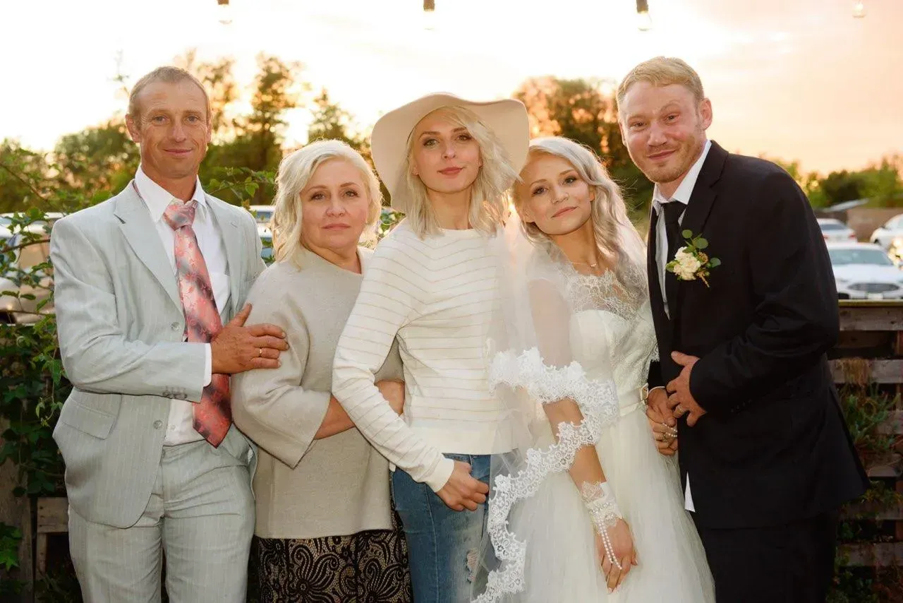 Family posing outdoors: bride in white dress, parents, siblings. Evening light.