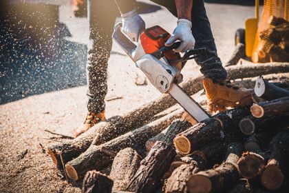 Person in jeans and gloves uses a chainsaw to cut firewood outdoors, sawdust flying.