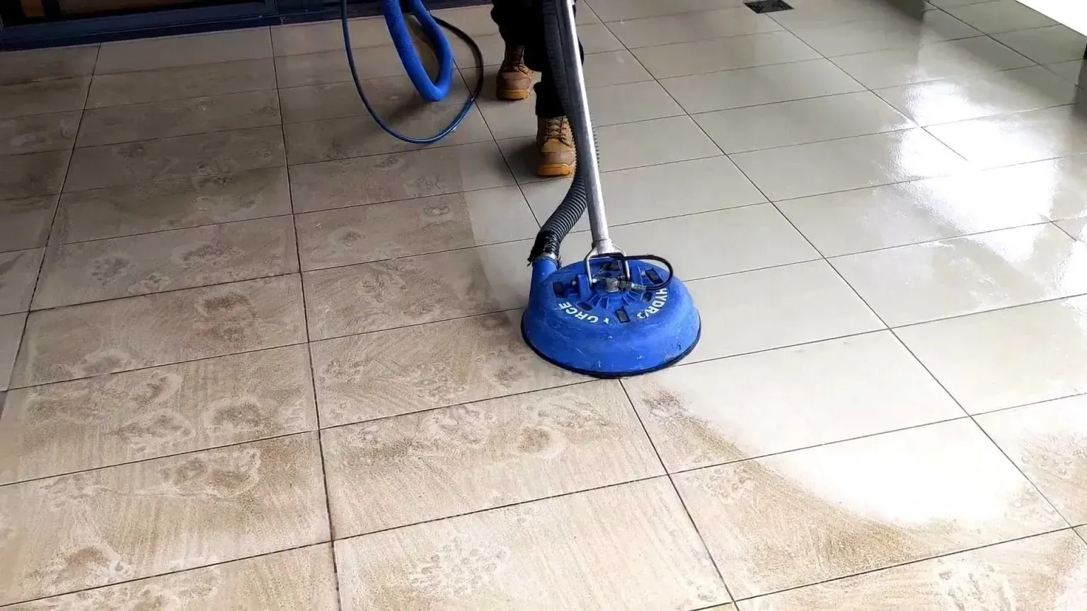 A person cleaning tile floor with a blue circular cleaning machine. Wet tile, some clean, some dirty.