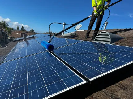 Person cleaning solar panels on a rooftop with a long brush and water. Blue sky.