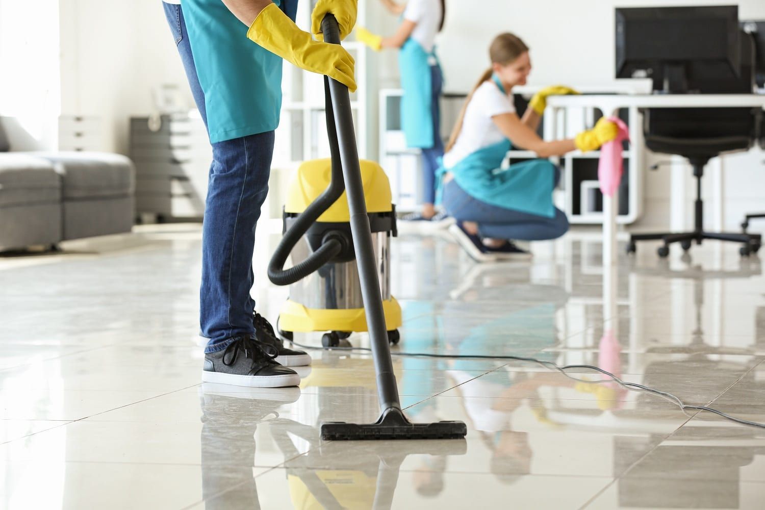 Three people cleaning an office. One vacuums, two dust furniture; all wear aprons and gloves.
