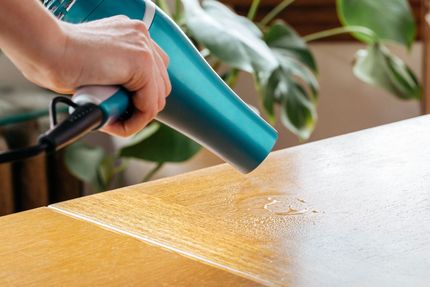 Hand holding a teal hair dryer, drying water from a light wood table surface.