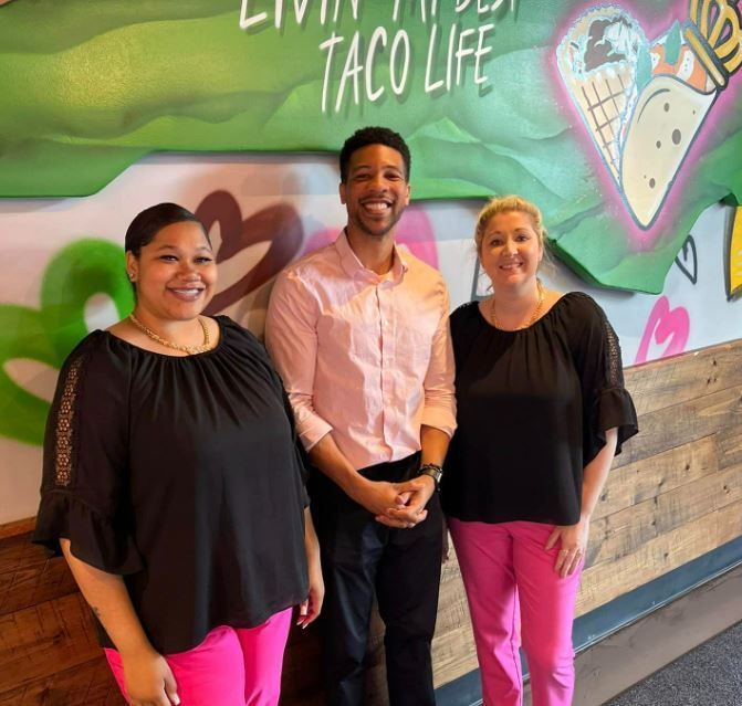 Three people standing in front of a wall that says taco life