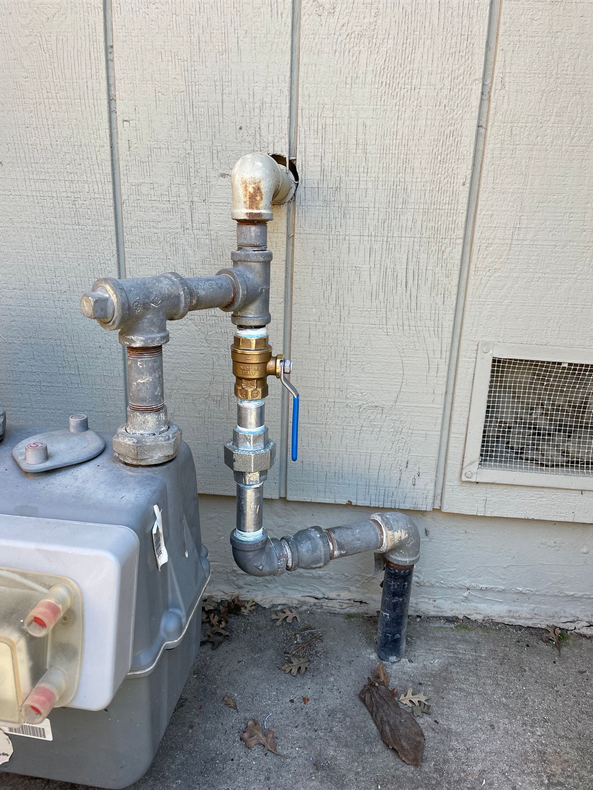 A gray residential gas meter connected to pipes extending from a building exterior wall.