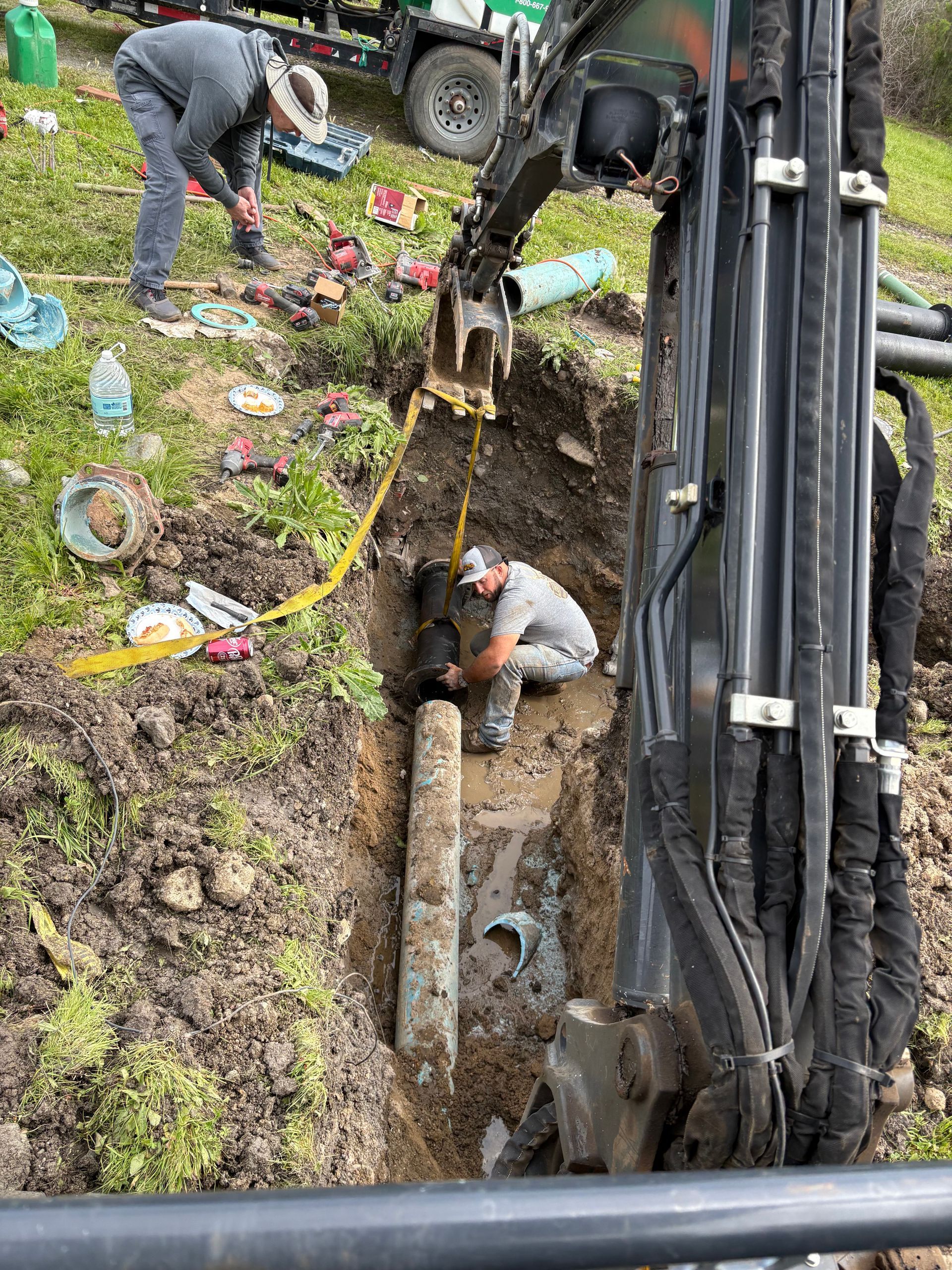 A worker kneels in a trench to repair a pipe, while a second person stands nearby next to an excavator.