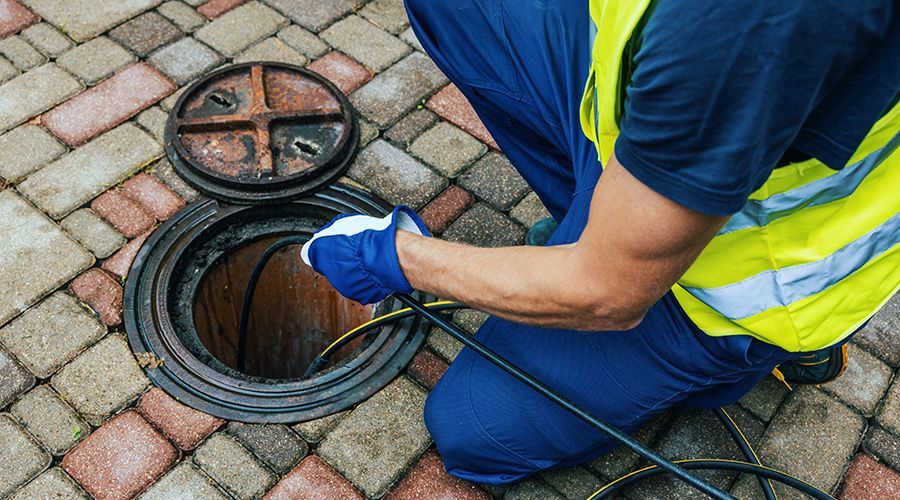 A worker in a high-visibility vest and blue uniform uses a sewer camera inspection cable to probe an open manhole cover.