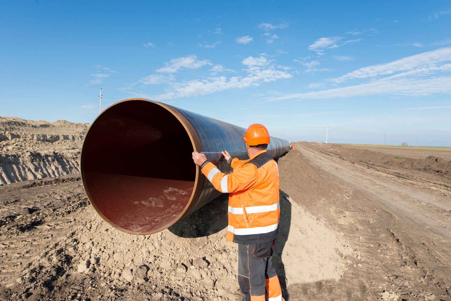A person in a high-visibility orange jacket and hard hat stands next to a large industrial pipe on a construction site.