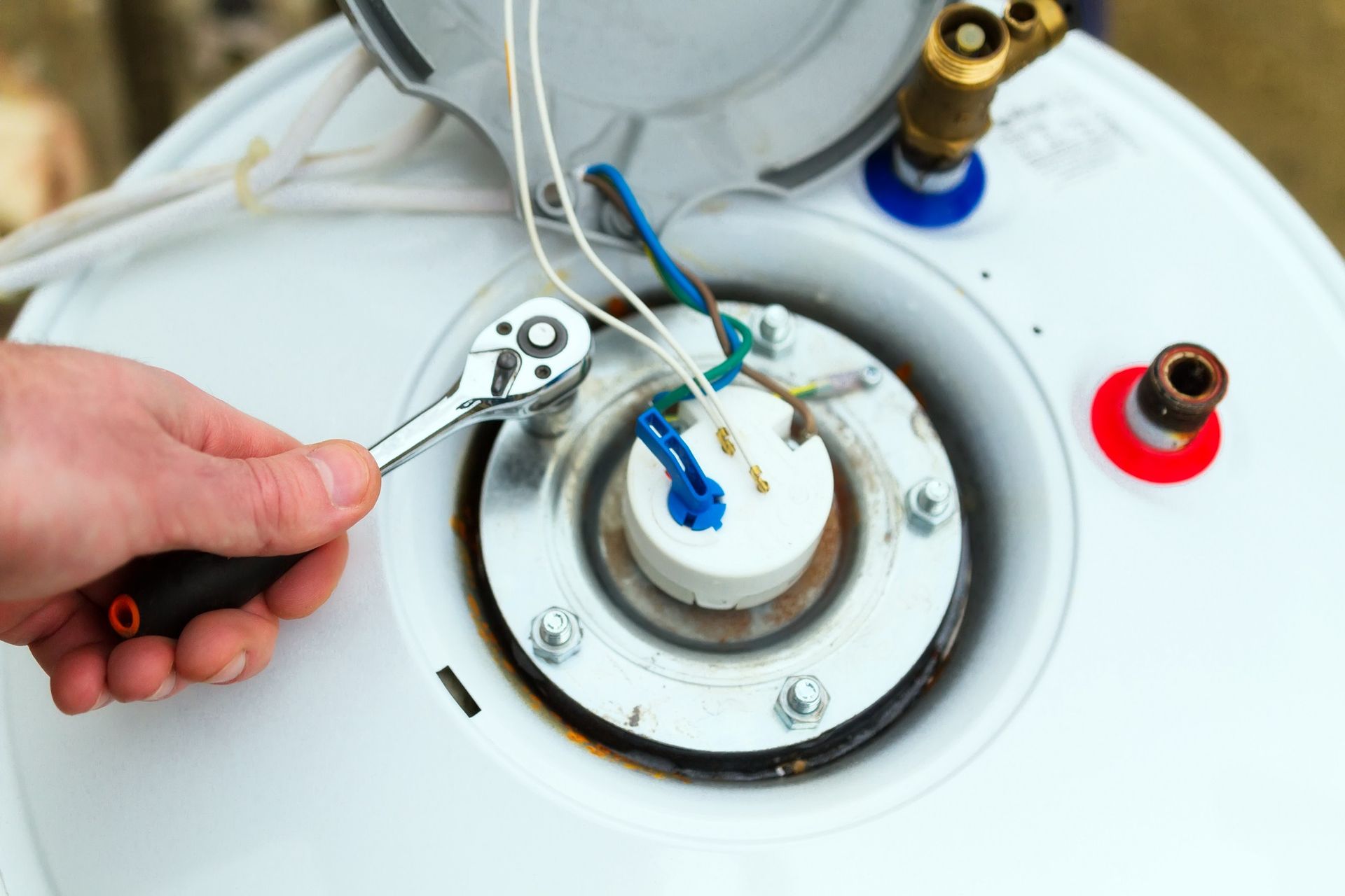 A hand uses a ratchet tool to tighten a bolt on an electric water heater's heating element assembly.