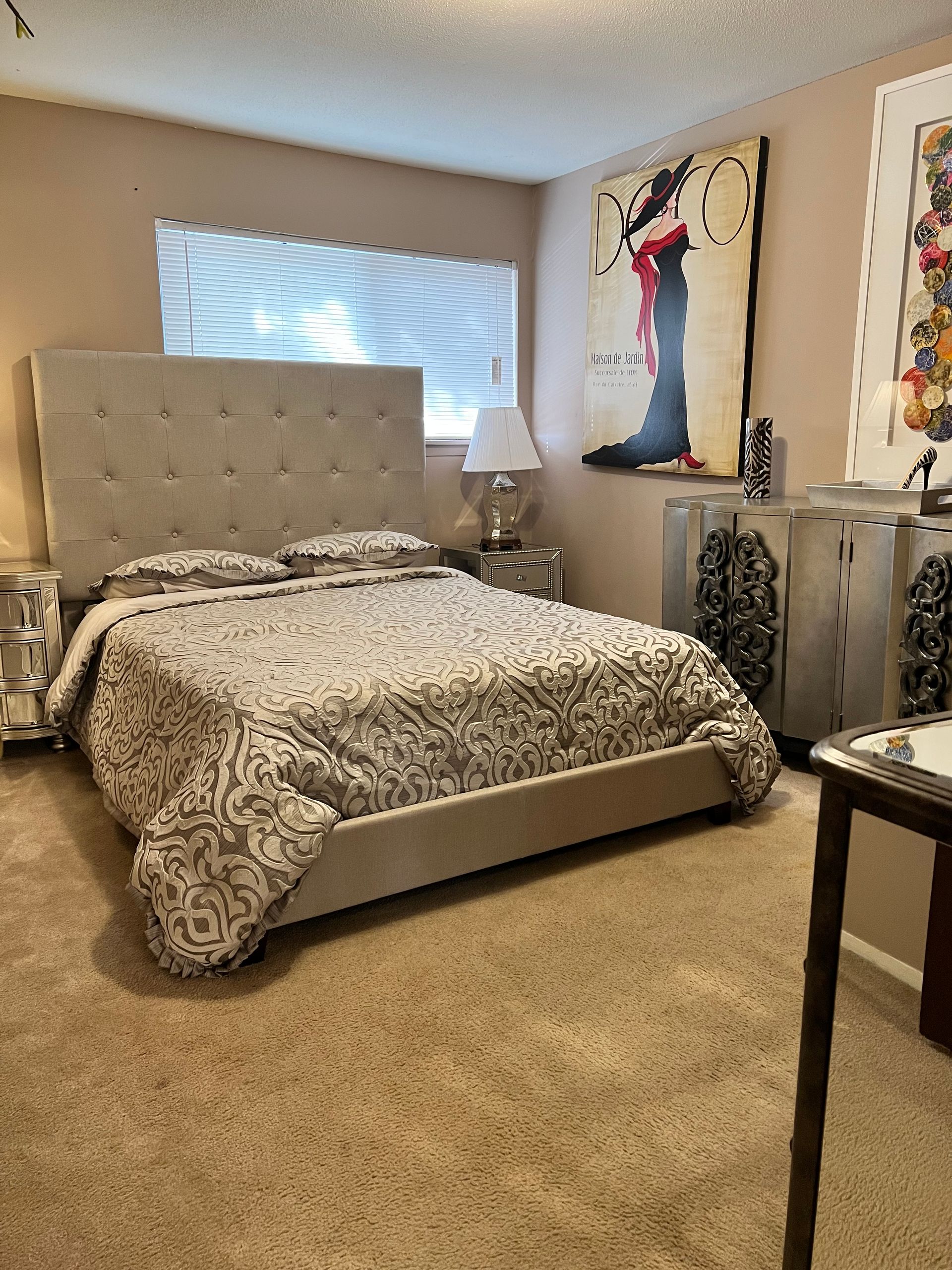 Bedroom with a bed, nightstands, artwork, and a window; tan walls and carpet.