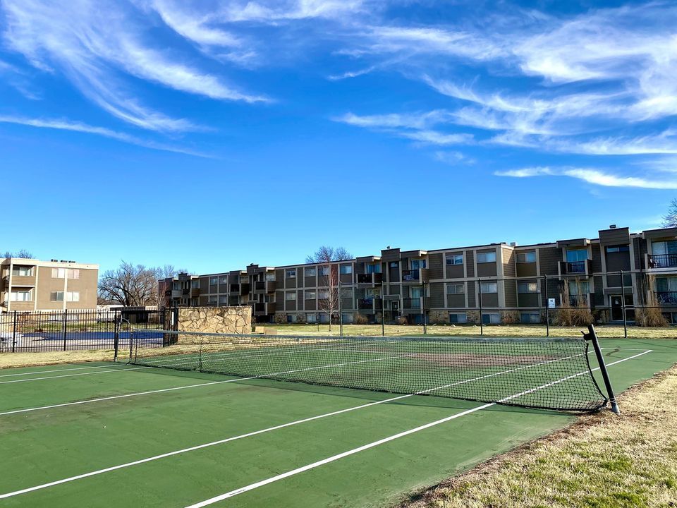 Green tennis court with net, buildings in background, blue sky with wispy clouds.