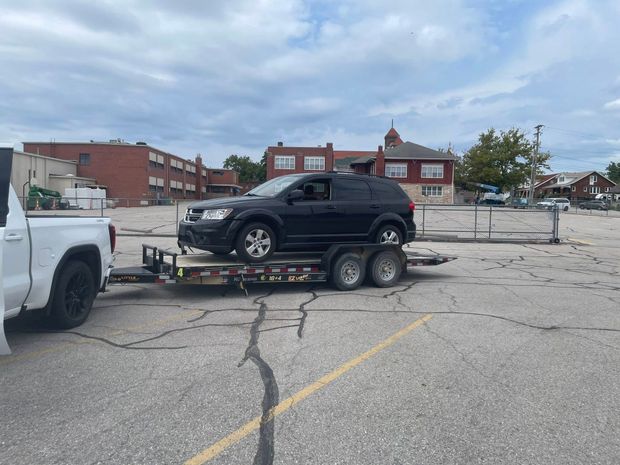 Black SUV on a trailer towed by a white truck in a parking lot near a brick building.