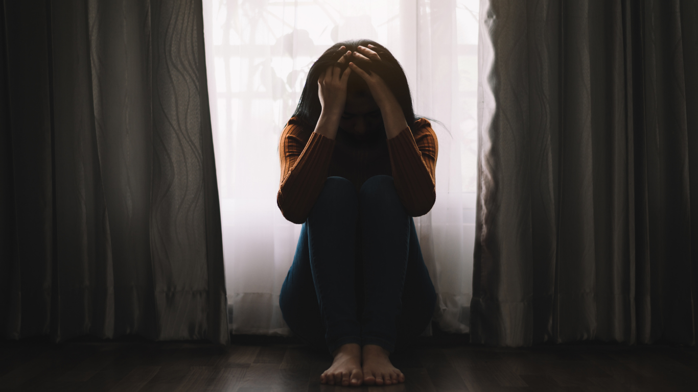 Woman sitting, head in hands, by a window with curtains. Dark, somber setting.