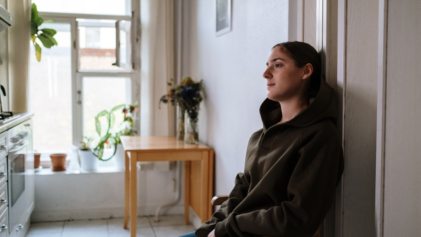 Woman in olive jacket leans against a white wall in a sunlit room, gazing toward window.