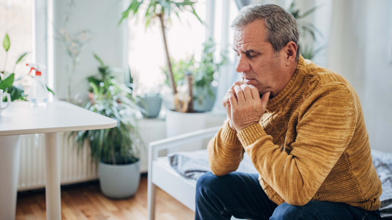Older man with gray hair in yellow sweater, looking down pensively, indoors.