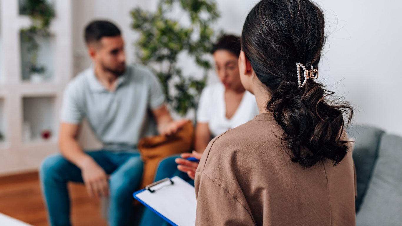 A therapist talking to a couple in a counseling session; the focus is on the therapist with a clipboard.