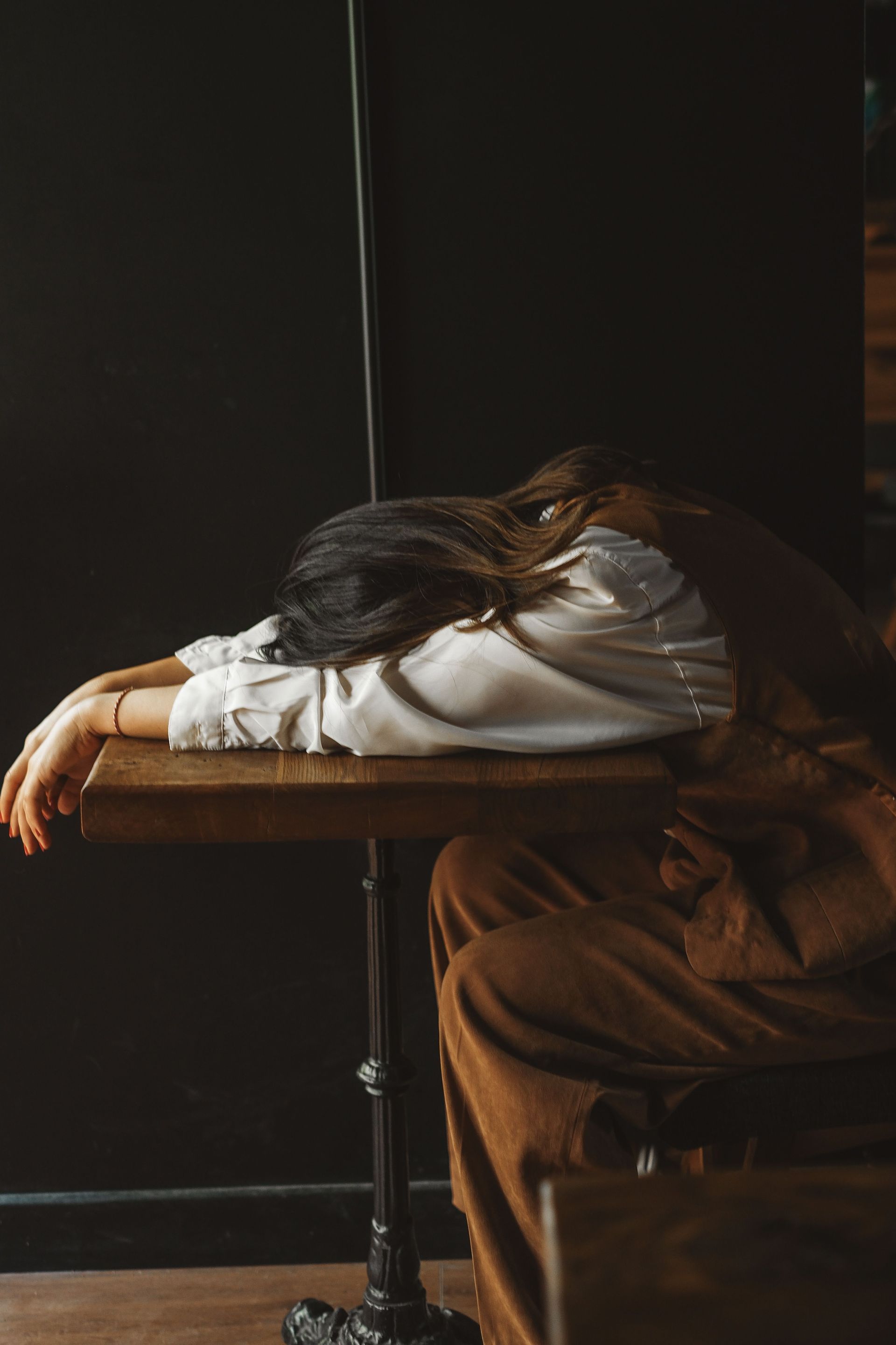 Person resting face down on a table in a dim room, wearing a white shirt and brown pants.