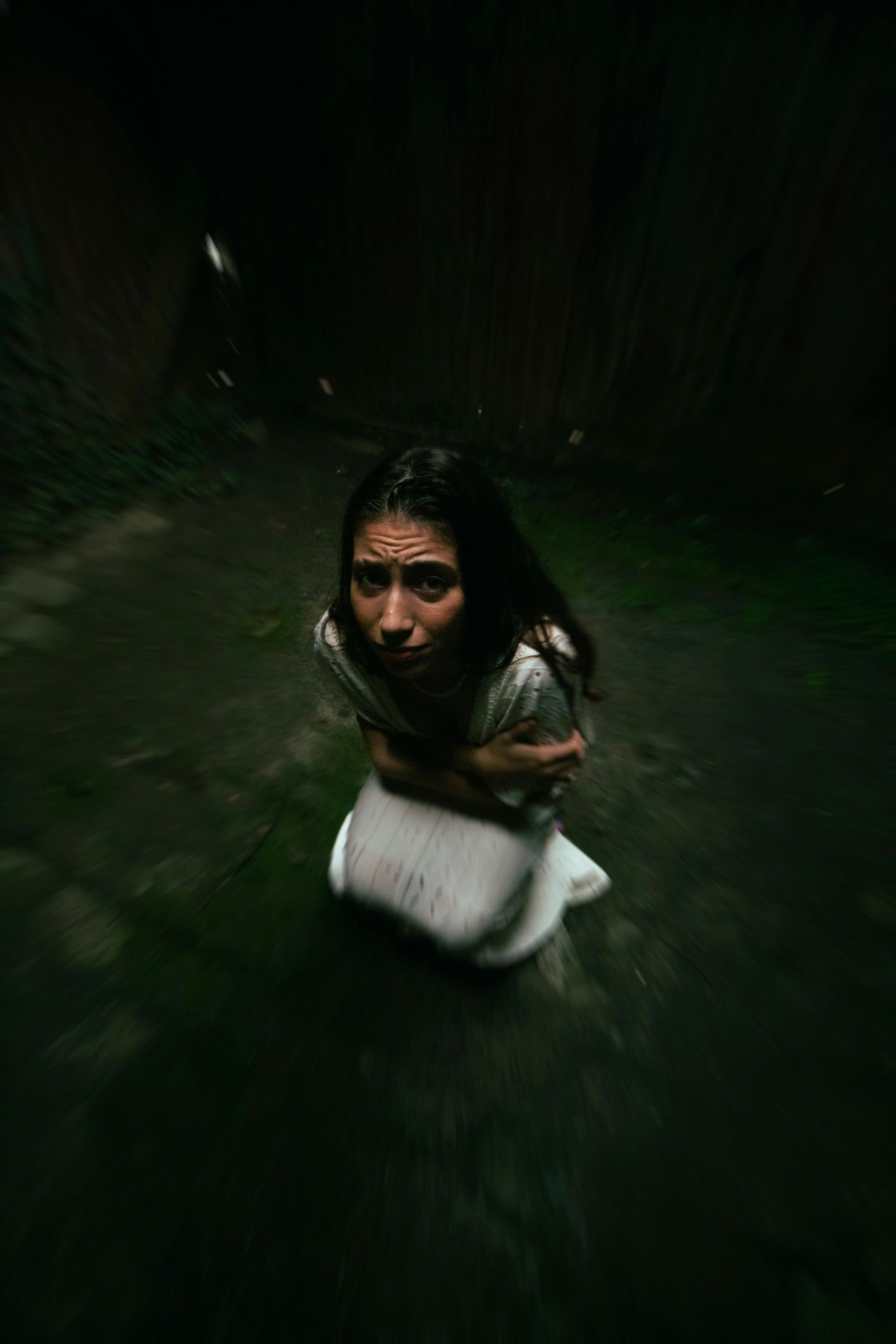A person crouched in the dark, wearing a white garment, looking upward with a somber expression in a dimly lit, moody space.