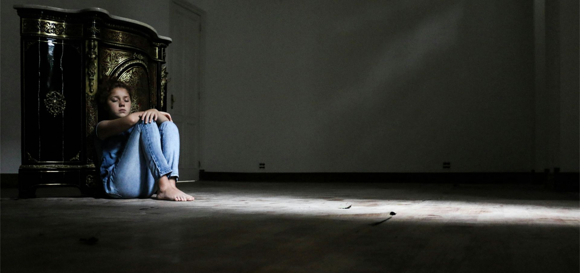 A person sitting on a dark floor in jeans, knees pulled to chest, beside an ornate wooden cabinet in a dimly lit room.