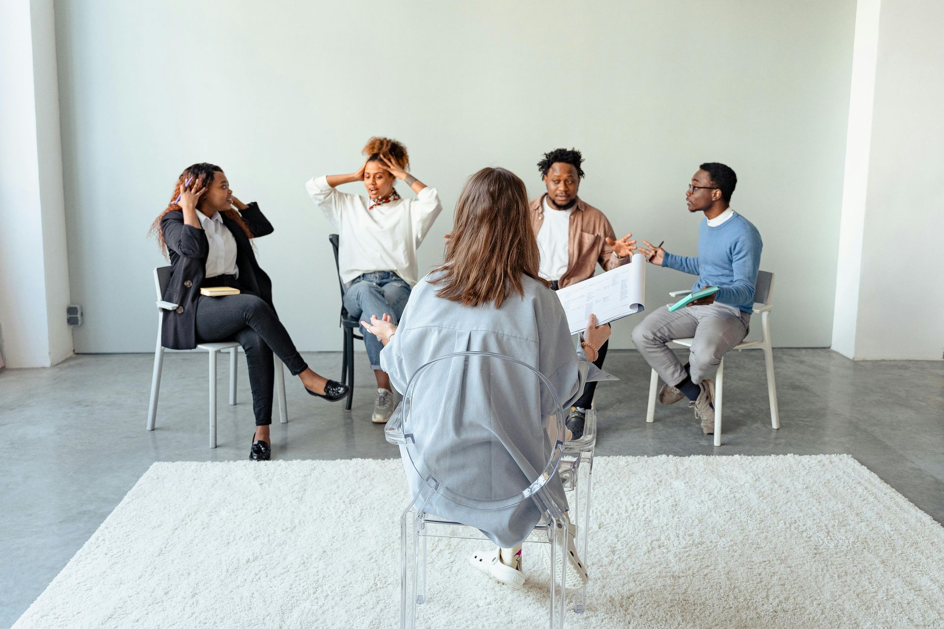 Group therapy session: People seated in a circle, discussing with therapist, one person holding head in hand.