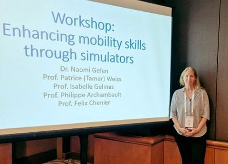 A woman is standing in front of a large screen that says workshop enhancing mobility skills through simulators.