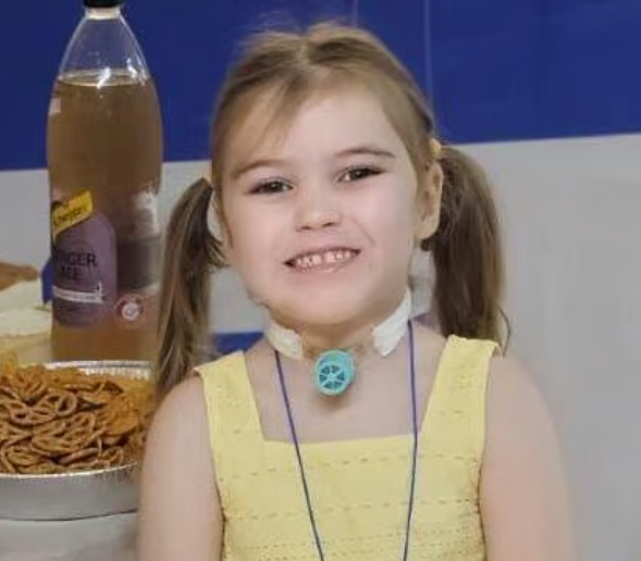 A little girl in a yellow dress is smiling in front of a bottle of schweppes