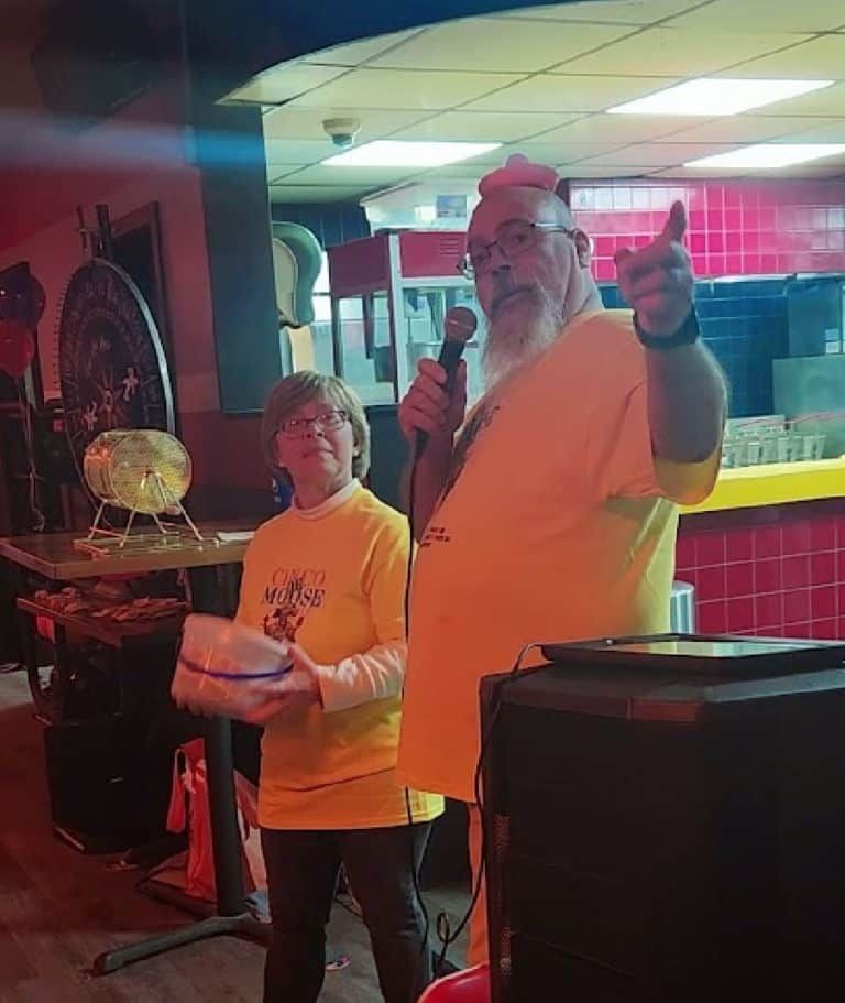 A man with a beard is holding a microphone while standing next to a woman in a yellow shirt.