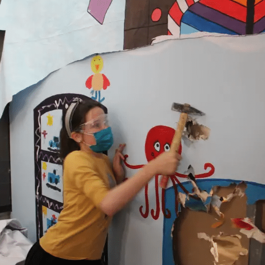 A woman wearing a mask is holding a hammer in front of a wall with an octopus on it
