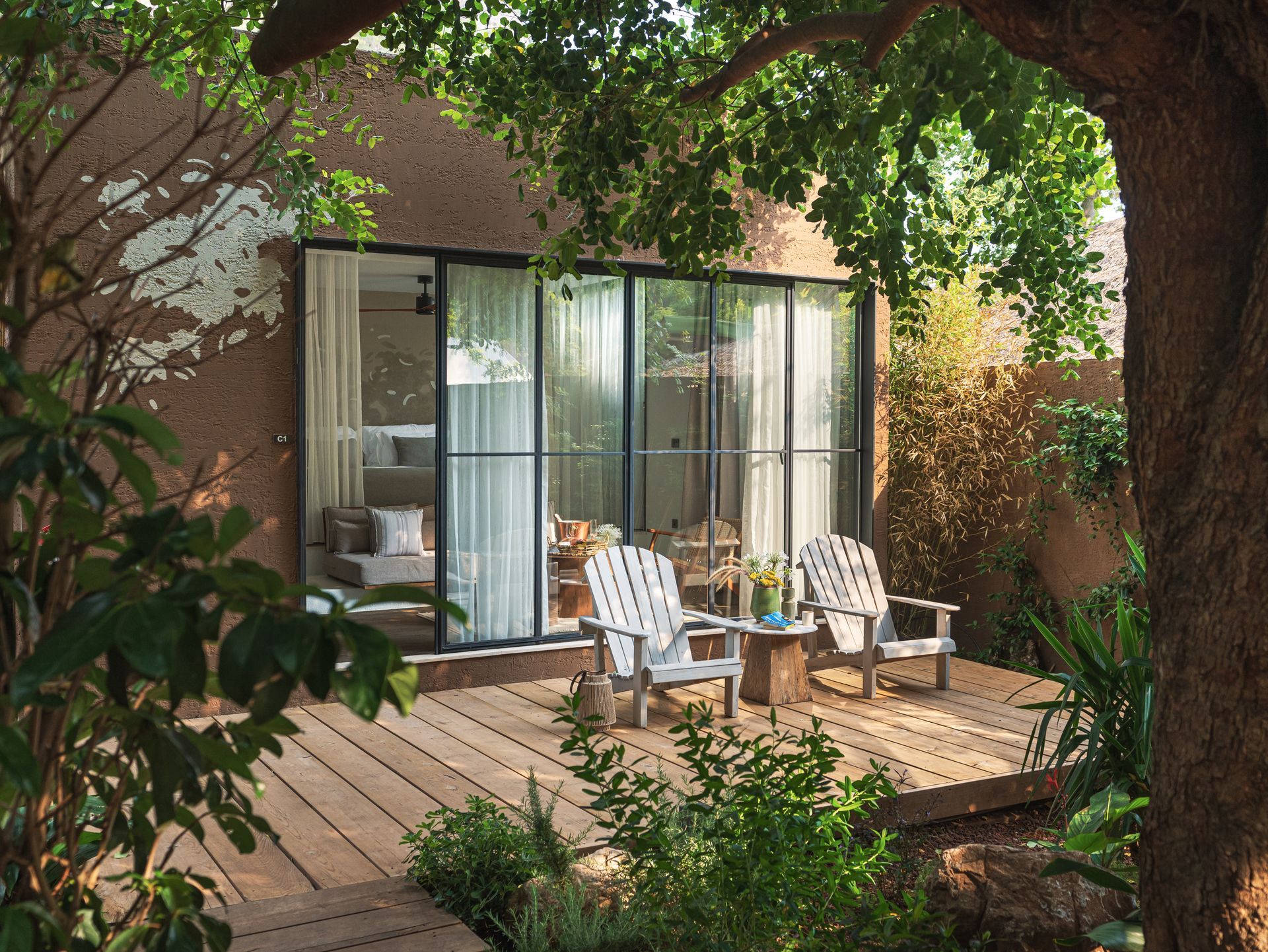 Two white chairs on a wooden deck outside a glass-walled room, surrounded by lush green trees and plants.
